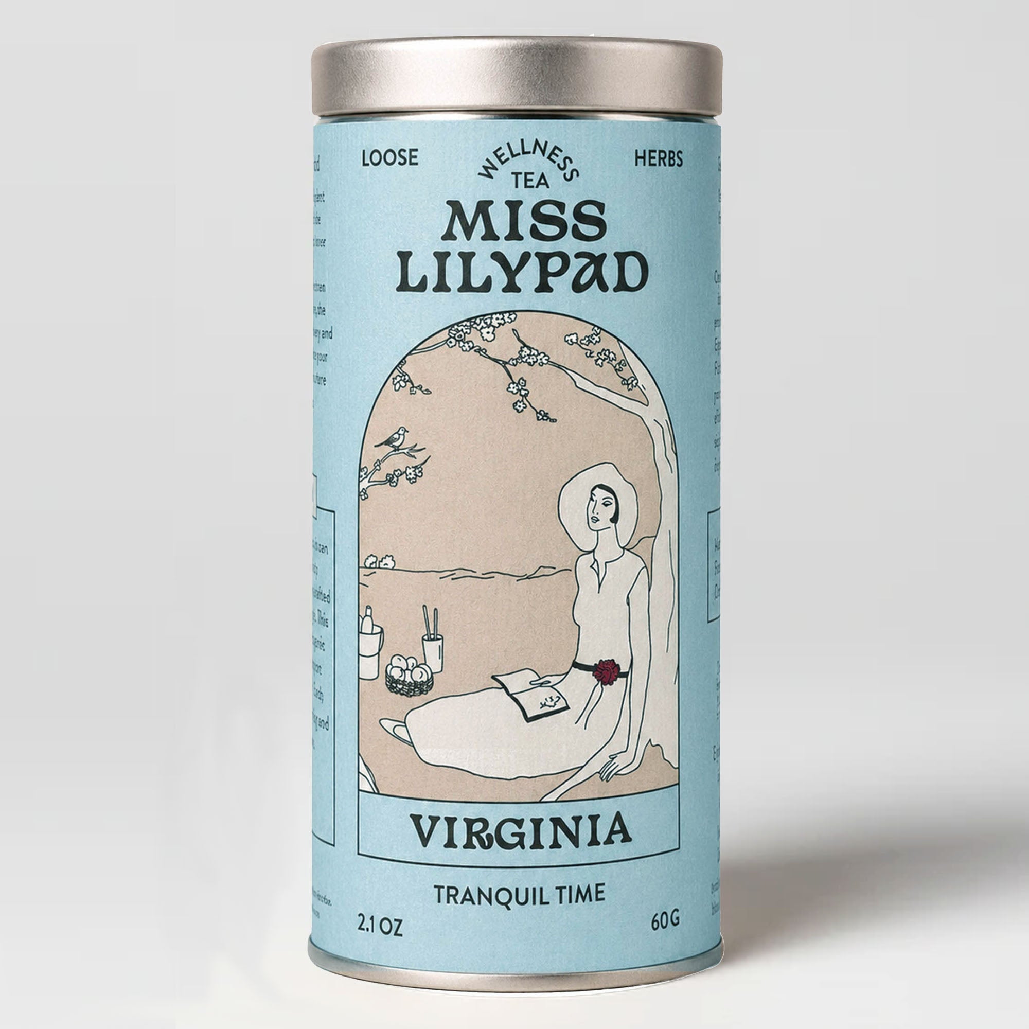 Miss Lilypad Tranquil Time Virginia loose herbal tea in a cylindrical tin canister with a blue label and silver metal lid and base. An illustration of a woman sitting under a tree reading a book wearing a red flower  belt